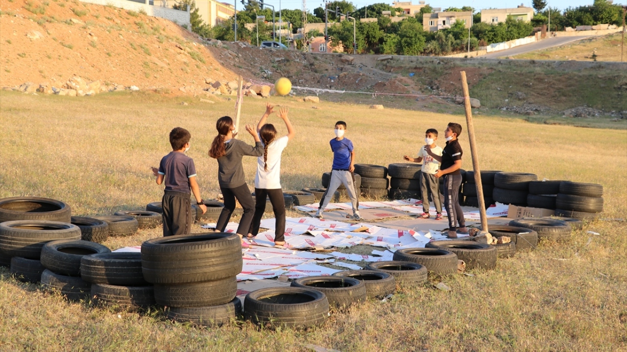 Sahaya karton döşeyerek düşme anında dikenlerden korunmaya çalışan çocuklar, alanda sosyal mesafe kuralına uyarak ve maske takarak voleybol oynuyor, eğlenceli vakit geçiriyor.